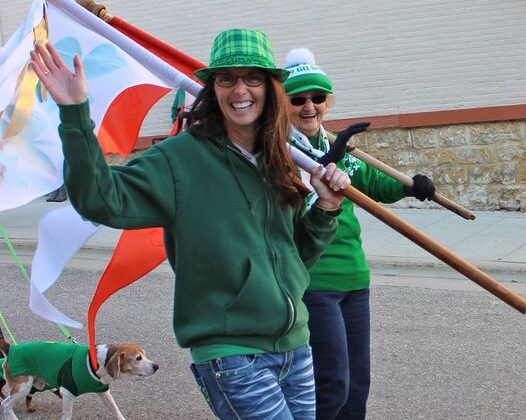 Two adults and a dog wearing green waving from a parade